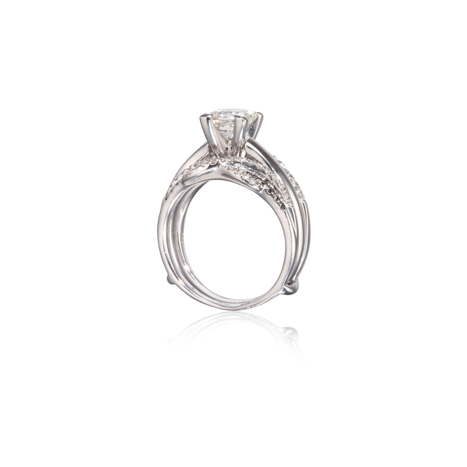 A white gold solitaire engagement ring with a round-cut center stone and small accent stones along the band, shown on a clean white background with a soft reflection below.