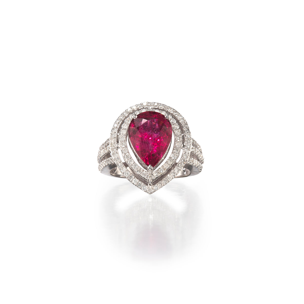 A pear-cut ruby ring set in white gold metal with a double halo of small clear stones, shown on a clean white background with a soft reflection below.