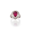 A pear-cut ruby ring set in white gold metal with a double halo of small clear stones, shown on a clean white background with a soft reflection below.