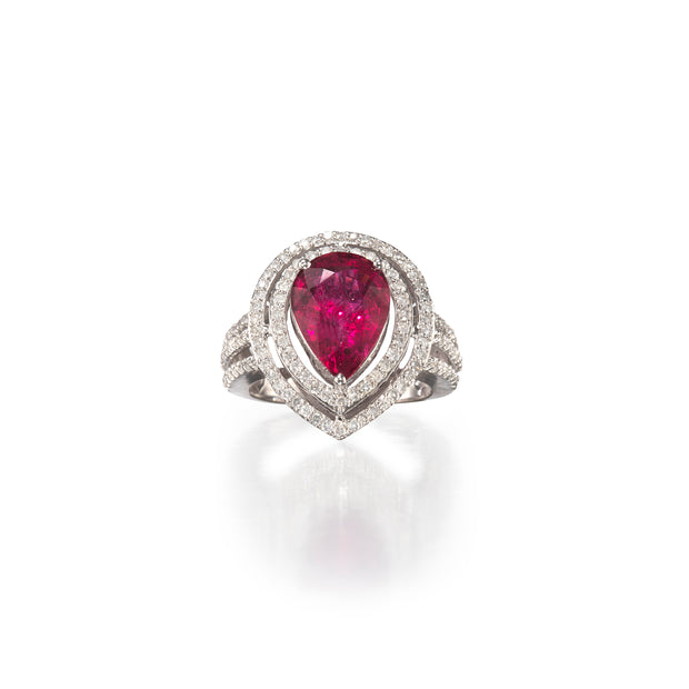 A pear-cut ruby ring set in white gold metal with a double halo of small clear stones, shown on a clean white background with a soft reflection below.