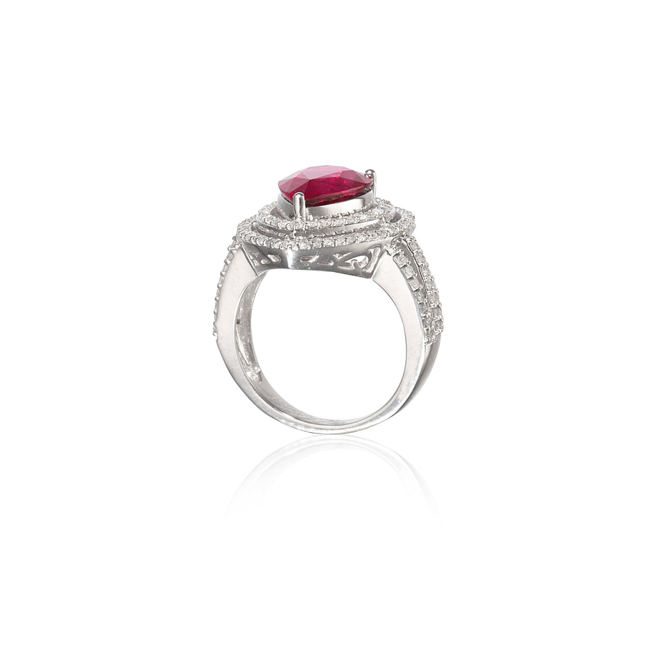 A white gold ring featuring a prominent oval-cut red gemstone in a prong setting, surrounded by a halo of small clear stones and an openwork patterned band, shown on a white background with a soft reflection.