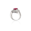 A white gold ring featuring a prominent oval-cut red gemstone in a prong setting, surrounded by a halo of small clear stones and an openwork patterned band, shown on a white background with a soft reflection.