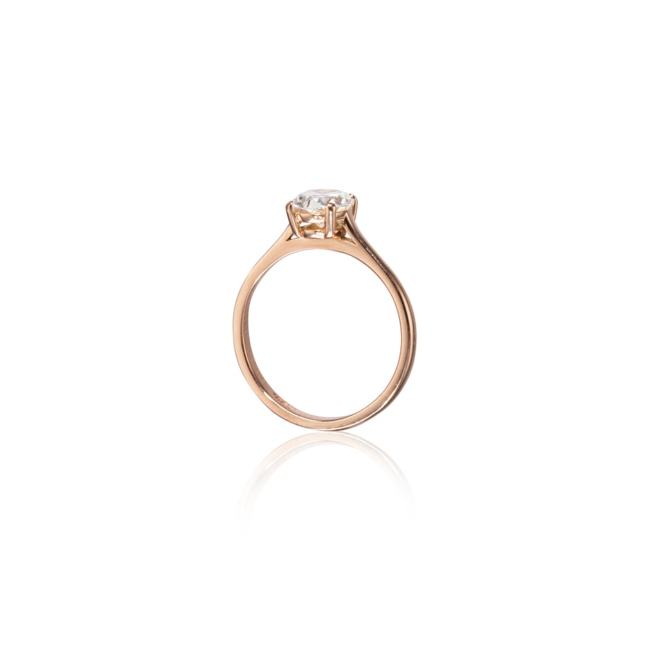 A rose gold solitaire ring with a round-cut clear gemstone in a four-prong setting, shown on a clean white background with a soft reflection below.