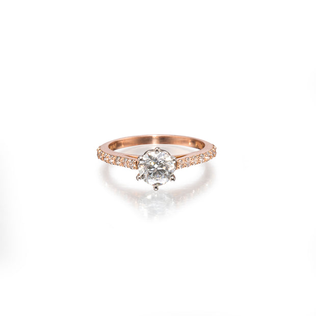 A rose gold engagement ring featuring a round-cut center stone in a prong setting, with a pavé band accented by small round stones, shown on a clean white background with a soft reflection.
