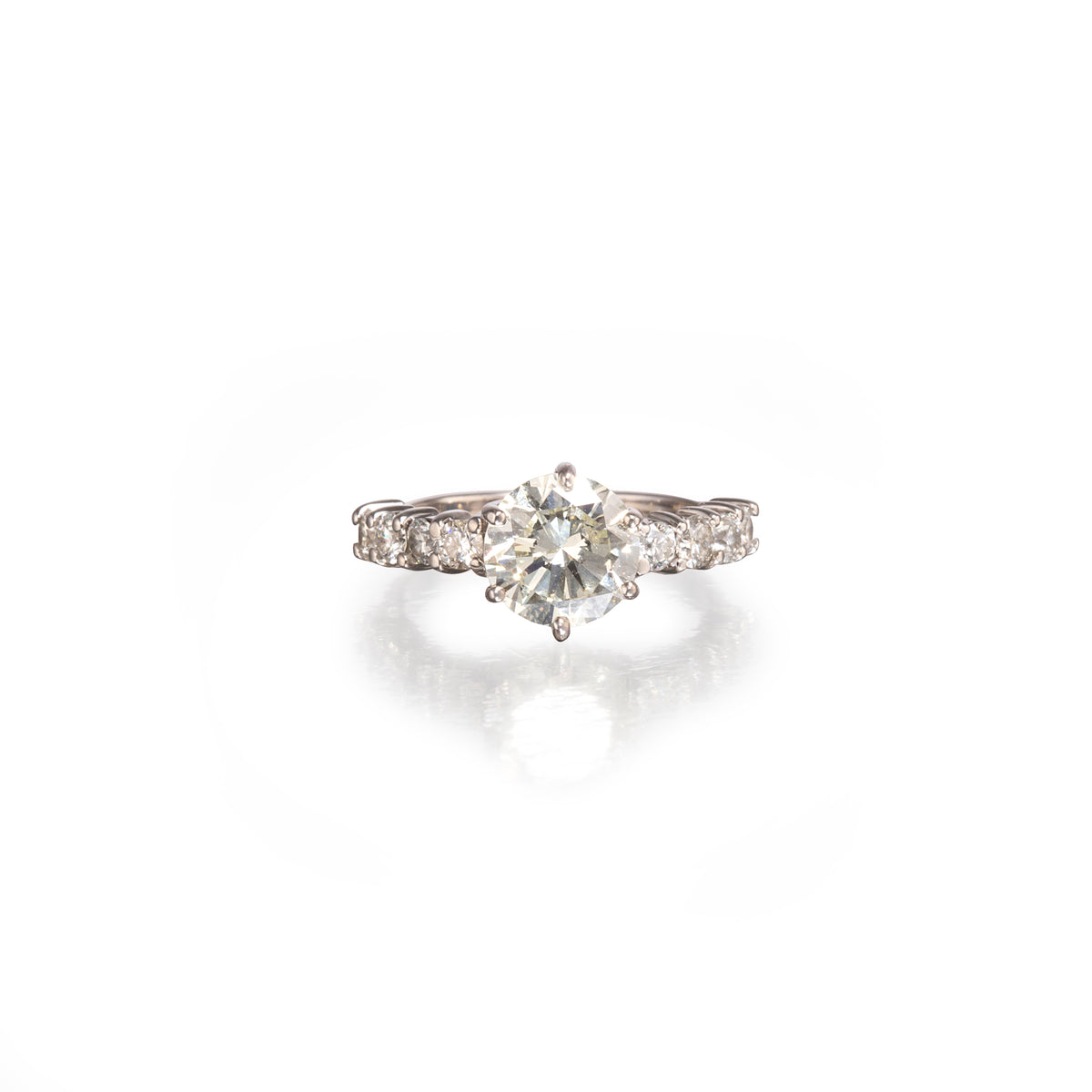 A white gold diamond ring with a round-cut center stone and smaller diamonds along the band, shown on a clean white background with a soft reflection below.