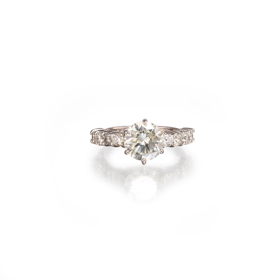 A white gold diamond ring with a round-cut center stone and smaller diamonds along the band, shown on a clean white background with a soft reflection below.