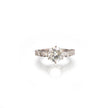 A white gold diamond ring with a round-cut center stone and smaller diamonds along the band, shown on a clean white background with a soft reflection below.
