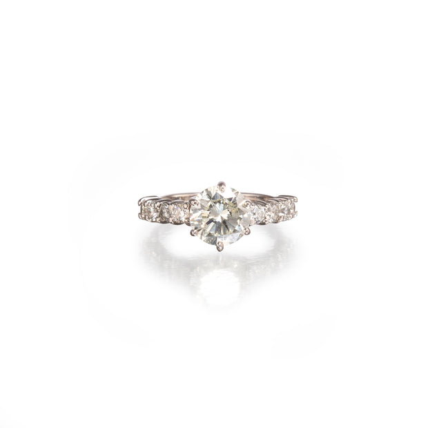 A white gold diamond ring with a round-cut center stone and smaller diamonds along the band, shown on a clean white background with a soft reflection below.