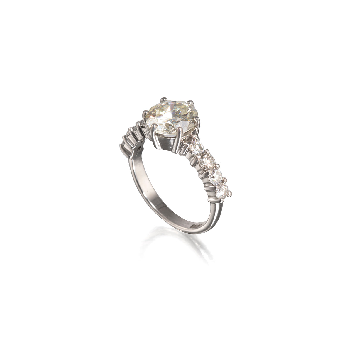 A white gold ring featuring a large round-cut clear center stone and a row of smaller clear stones along the band, shown on a plain white background with a soft reflection below.