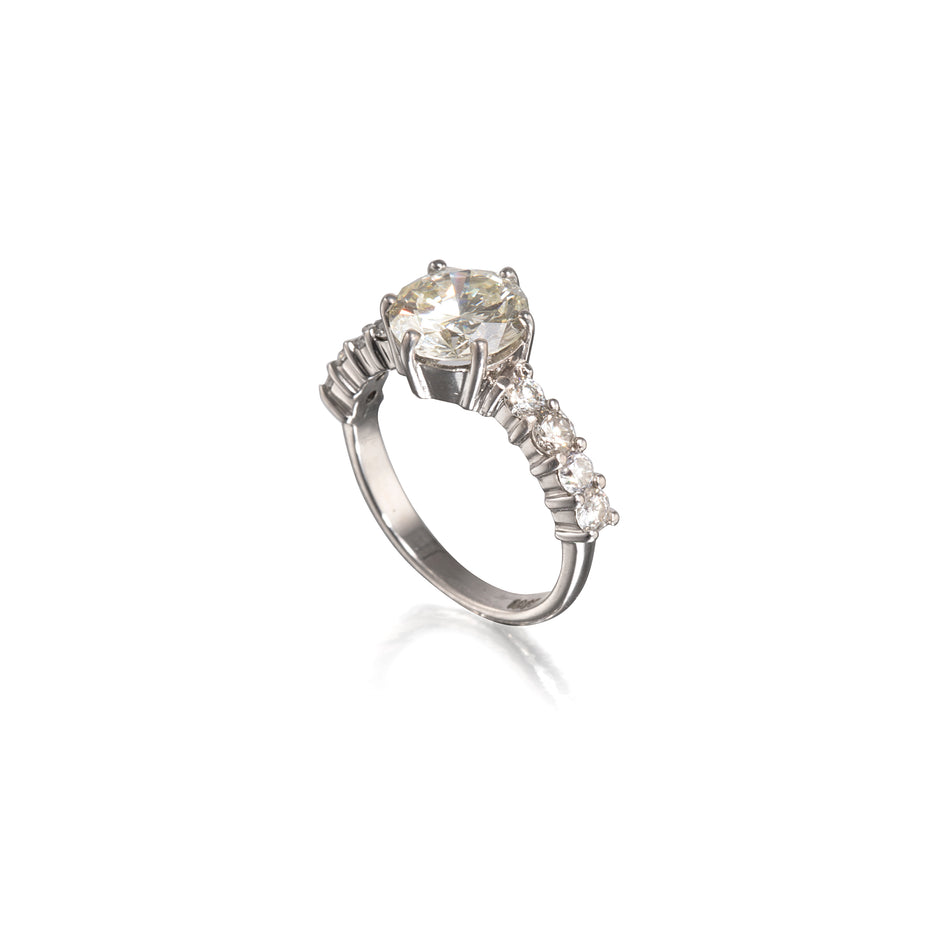 A white gold ring featuring a large round-cut clear center stone and a row of smaller clear stones along the band, shown on a plain white background with a soft reflection below.