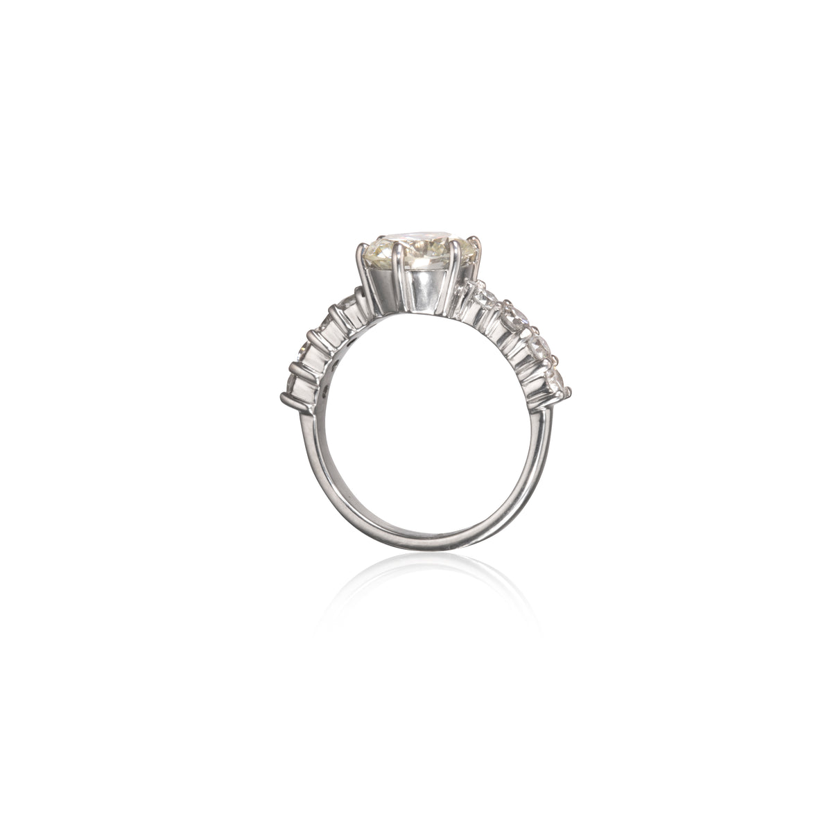 A white gold diamond ring with a large round center stone and a row of smaller baguette-cut diamonds on the shoulders, shown on a plain white background with a soft reflection below.
