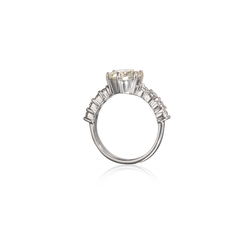 A white gold diamond ring with a large round center stone and a row of smaller baguette-cut diamonds on the shoulders, shown on a plain white background with a soft reflection below.
