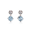 A pair of white gold drop earrings featuring a small clear round stone at the top and a larger light blue round stone below, set against a plain white background with a soft reflection.