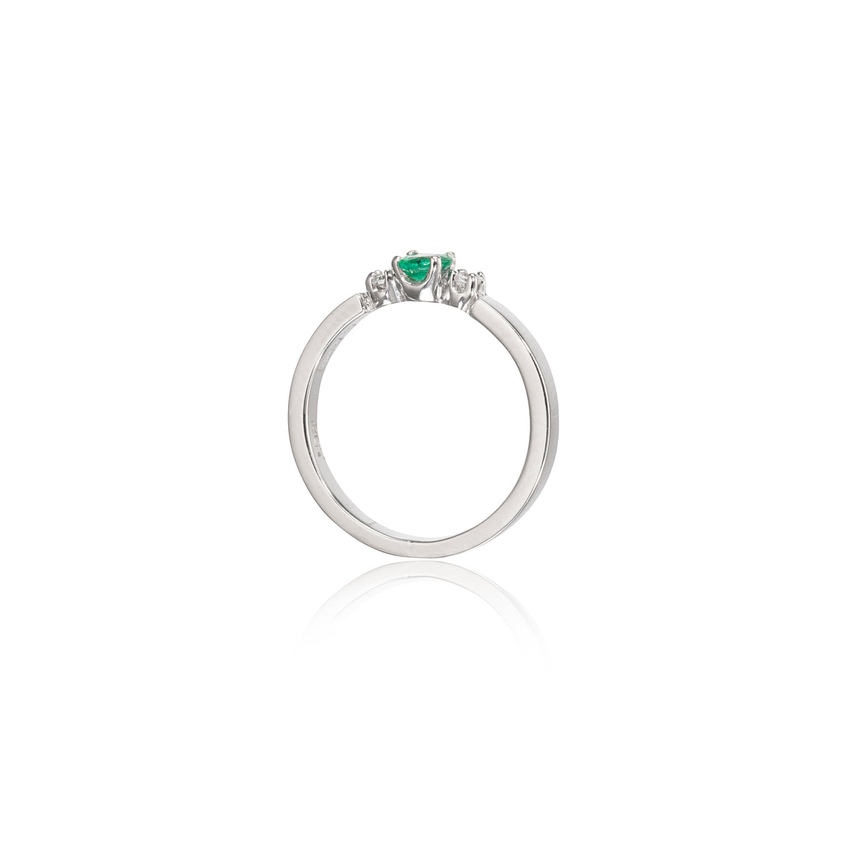 A white gold ring featuring a small green heart-shaped gemstone in the center, flanked by two tiny clear stones, shown on a white background with a soft reflection below.