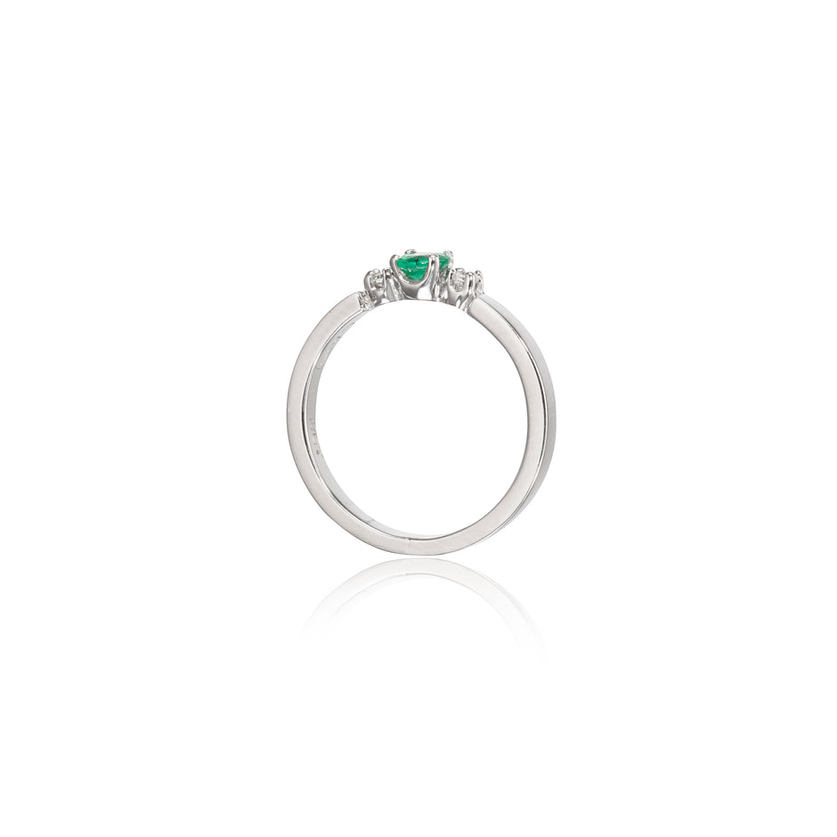 A white gold ring featuring a small green heart-shaped gemstone in the center, flanked by two tiny clear stones, shown on a white background with a soft reflection below.