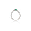 A white gold ring featuring a small green heart-shaped gemstone in the center, flanked by two tiny clear stones, shown on a white background with a soft reflection below.
