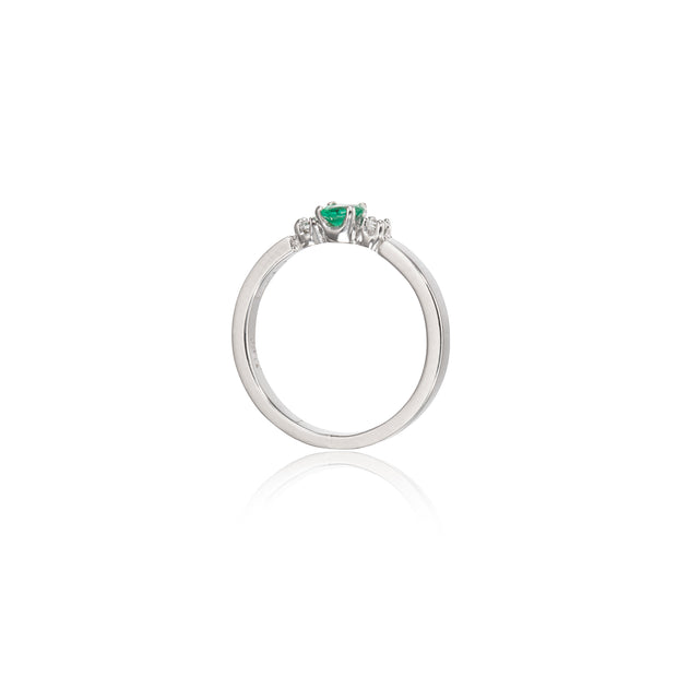 A white gold ring featuring a small green heart-shaped gemstone in the center, flanked by two tiny clear stones, shown on a white background with a soft reflection below.