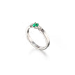 A white gold ring featuring a small oval-cut green emerald gemstone in a prong setting, accented by tiny clear stones on the shoulders, shown on a white background with a soft reflection.
