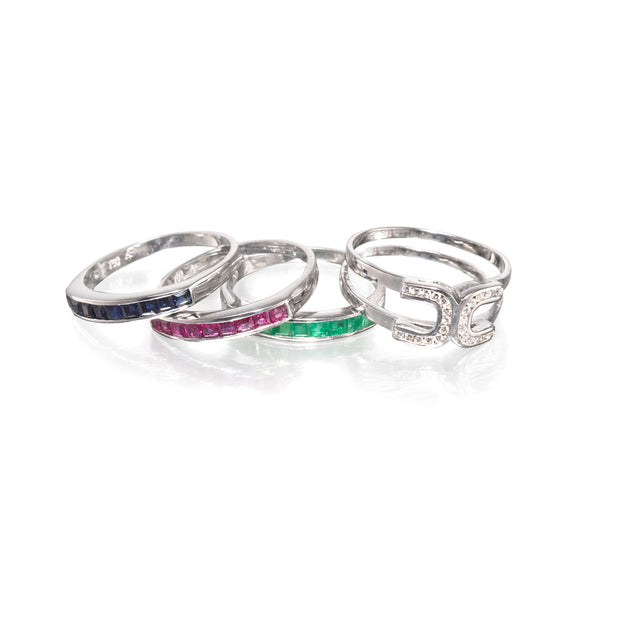 Four White gold rings arranged in a fan shape on a white reflective surface. Two rings are dark blue, one is pink, one is green, and one features clear stones.
