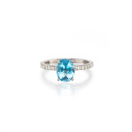 An oval-cut light blue topaz ring with a white gold  band paved with small clear stones, shown on a clean white background with a soft reflection.