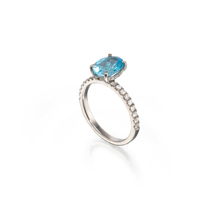A white gold  ring featuring a cushion-cut light blue gemstone in a prong setting, with a pavé band of small clear stones, shown on a clean white background.
