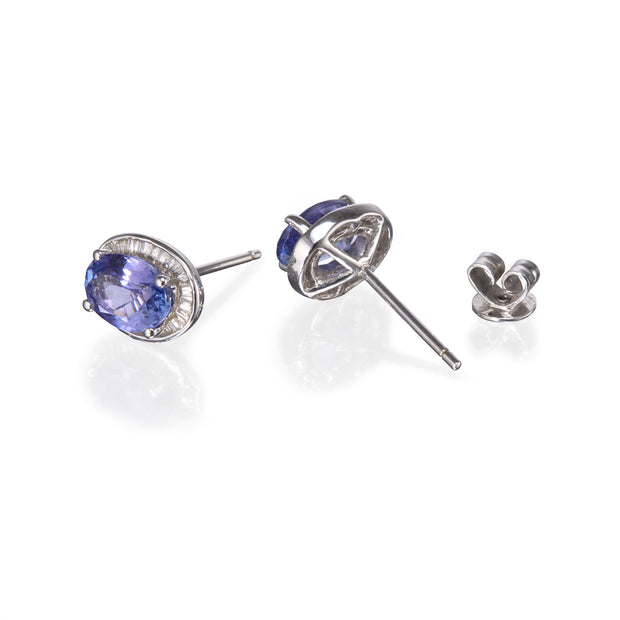A pair of oval tanzanite stud earrings in silver settings, shown with one earring facing forward and the other turned to show the post and butterfly back, on a white background.