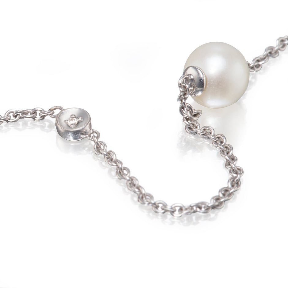 A close-up of a white gold chain necklace featuring a large, lustrous white pearl and a small round silver charm with a tiny clear stone, set against a clean white background.