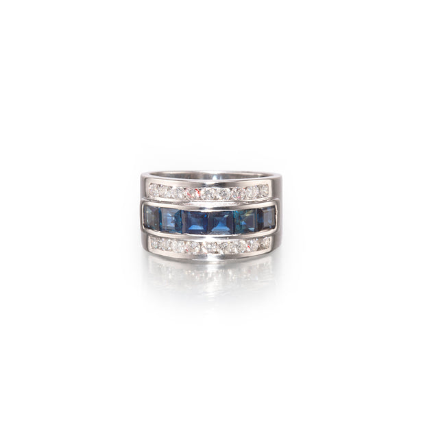 A white gold ring featuring three horizontal bands: the top and bottom bands are lined with small clear round stones, and the center band showcases five rectangular blue gemstones.