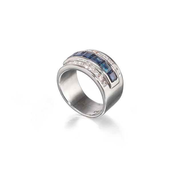 A white gold ring featuring a curved top set with dark blue rectangular stones on one side and small clear round stones on the other, photographed on a clean white background.