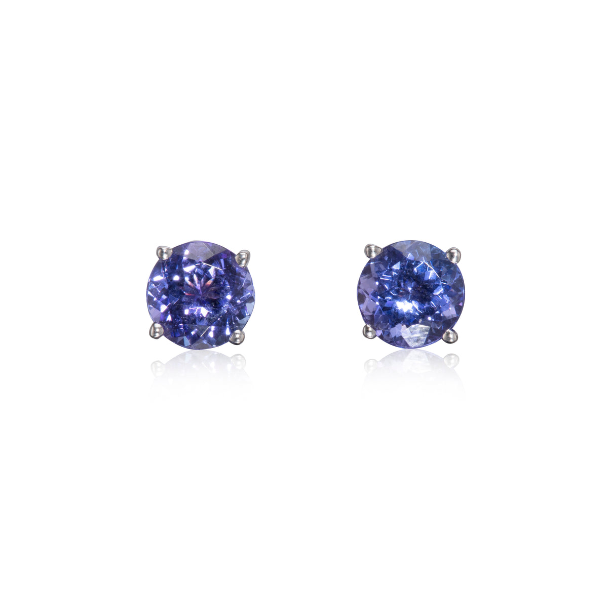 A pair of round-cut stud earrings featuring deep blue-purple tanzanite's in a four-prong setting, displayed on a clean white background with soft reflections below.