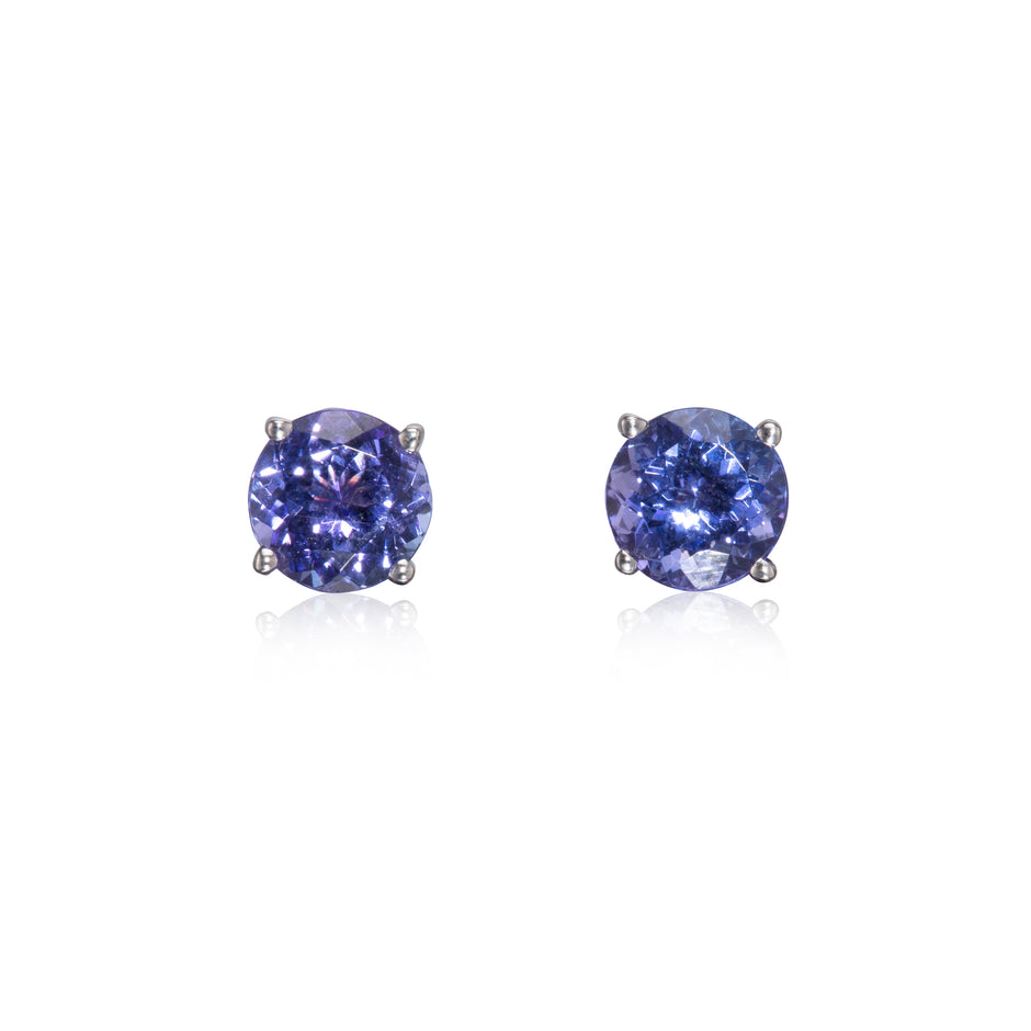 A pair of round-cut stud earrings featuring deep blue-purple tanzanite's in a four-prong setting, displayed on a clean white background with soft reflections below.