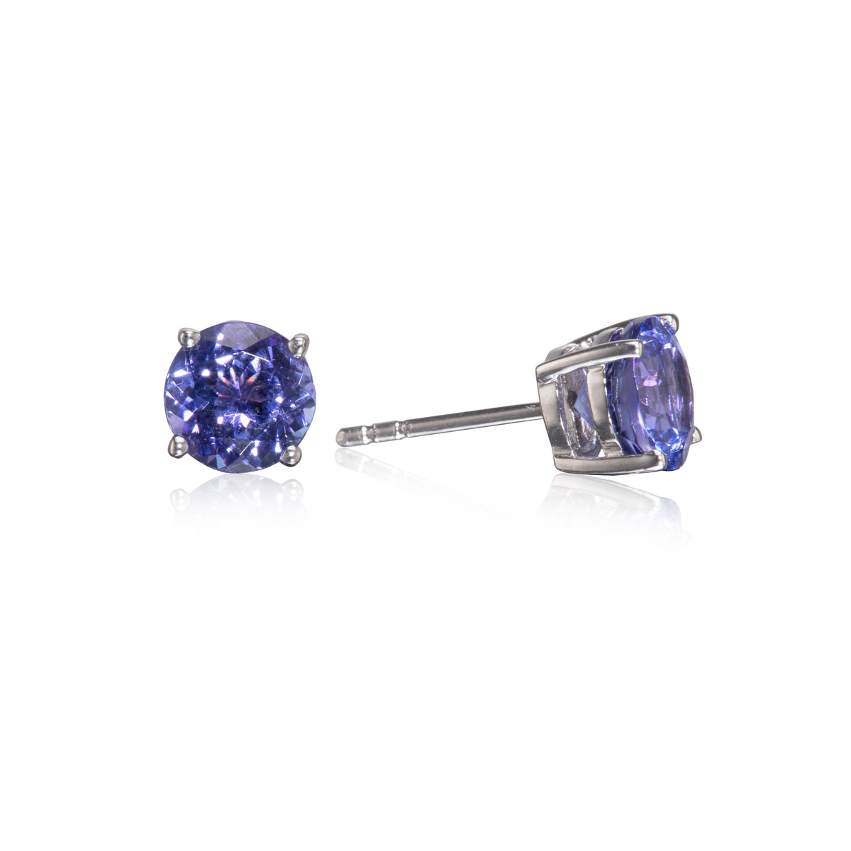 A pair of round-cut blue tanzanite stud earrings in silver four-prong settings, with one earring facing forward and the other angled to show the post and back, on a white background with a soft reflection.