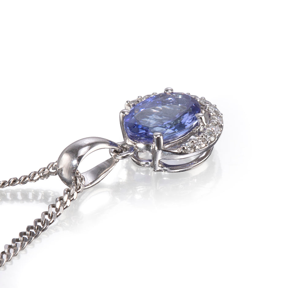 A white gold pendant necklace featuring a round blue tanzanite gemstone surrounded by a halo of small clear stones, shown on a white reflective surface.