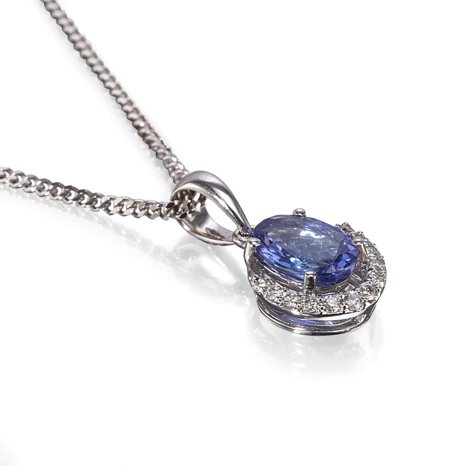 A white gold necklace with a halo pendant featuring a central oval blue tanzanite gemstone surrounded by small clear stones, on a white background.