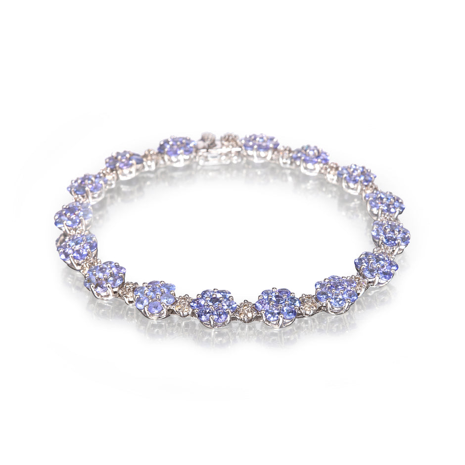 A white gold tennis bracelet featuring round light blue tanzanite gemstones and small clear accent stones, displayed on a clean white background with a soft reflection.