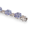 A close-up of a white gold tennis bracelet featuring round-cut light blue tanzanite gemstones in floral clusters, separated by small clear accent stones, on a white background.