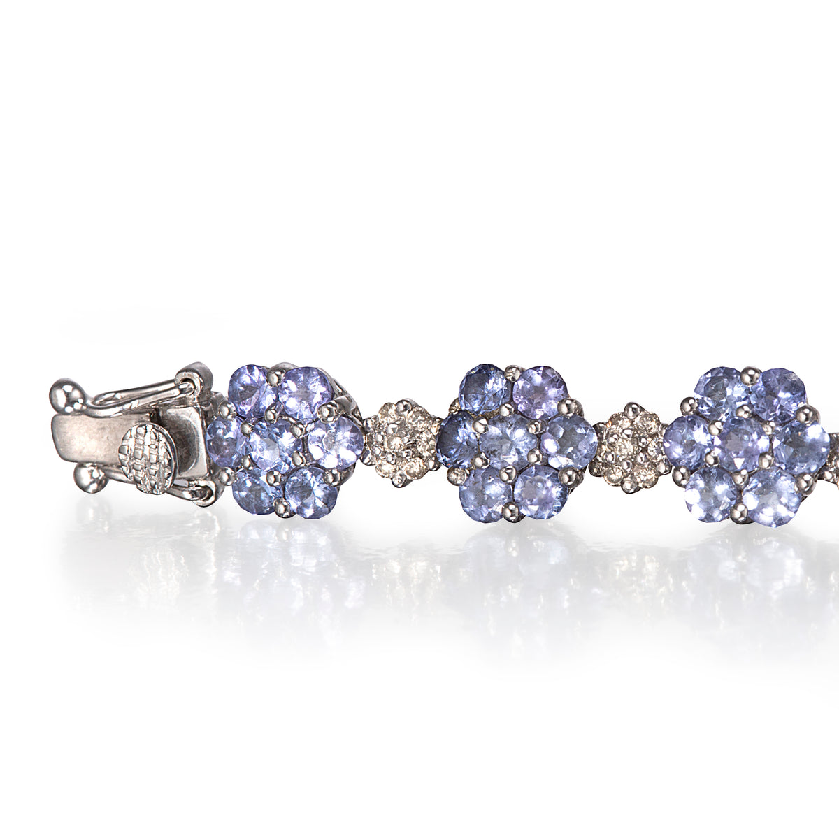A white gold tennis bracelet featuring three clusters of round-cut light blue tanzanite gemstones, separated by small diamond accents, shown on a reflective white surface.
