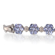 A white gold tennis bracelet featuring three clusters of round-cut light blue tanzanite gemstones, separated by small diamond accents, shown on a reflective white surface.