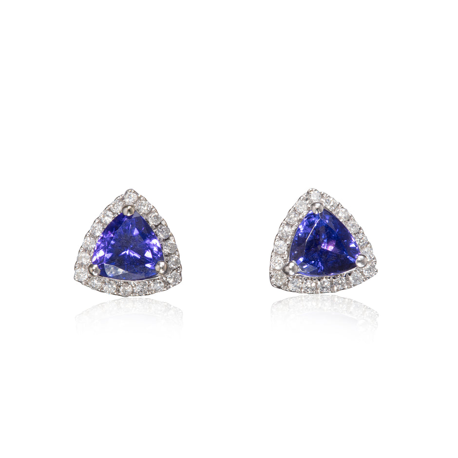 A pair of triangular stud earrings featuring a central blue tanzite gemstone surrounded by a halo of small clear stones in a white gold setting, shown on a white background with a soft reflection.