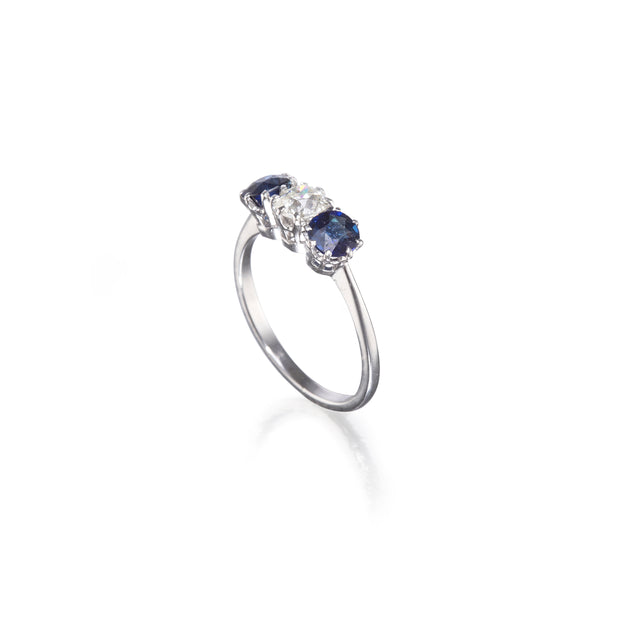 A white gold three-stone ring featuring a central round clear gemstone flanked by two oval deep blue sapphires, set against a clean white background with a soft reflection.