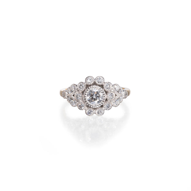 A white and yellow diamond ring with a central round diamond surrounded by a halo of smaller diamonds, set against a plain white background with a soft reflection below.
