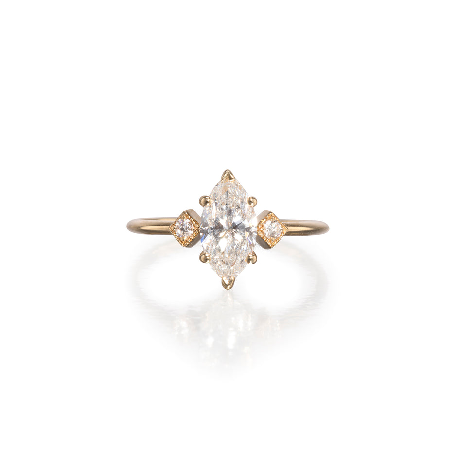 A yellow gold ring featuring a marquise-cut diamond center stone with two smaller round-cut diamonds on the sides, set against a clean white background with a soft reflection.
