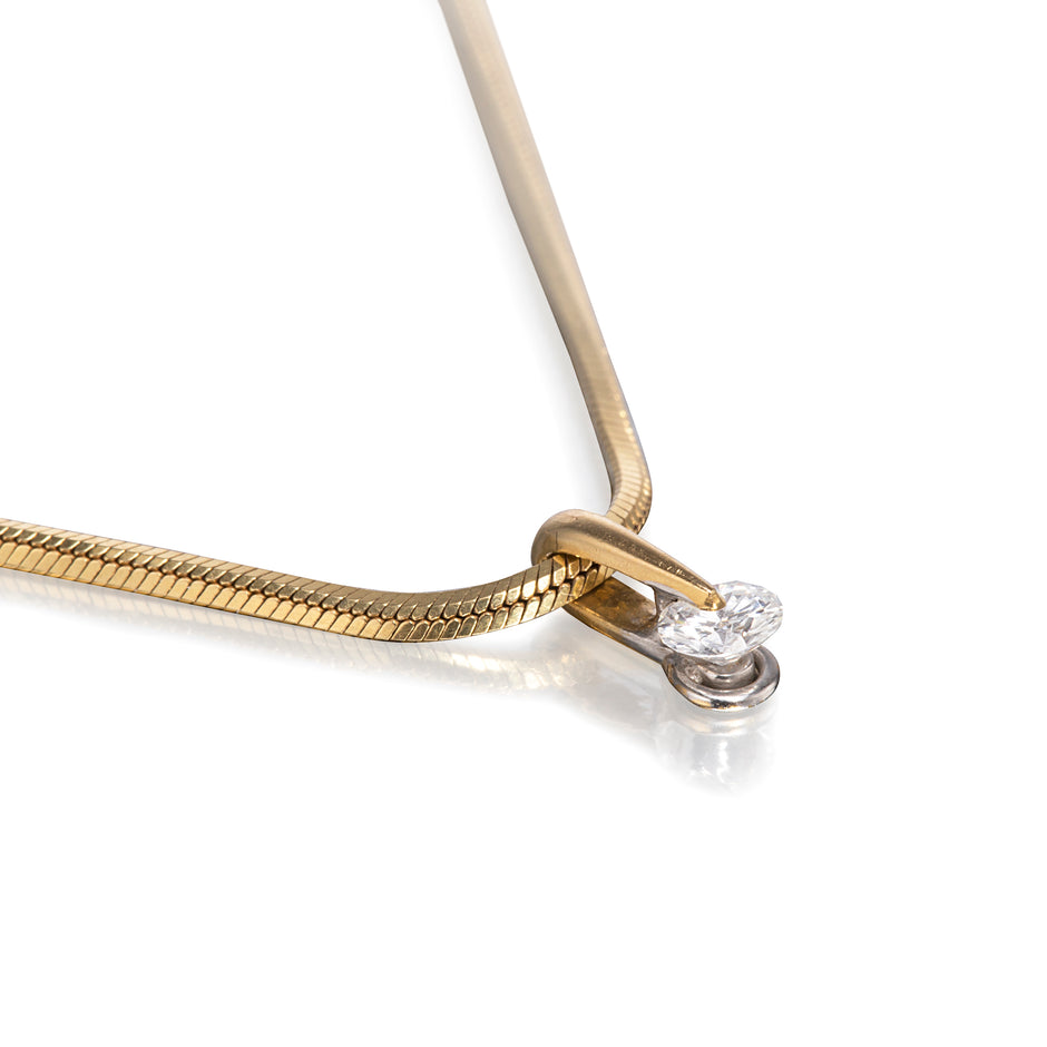 A close-up studio shot of a gold snake chain necklace with a small silver-toned setting holding two clear gemstones, photographed on a white background with a soft reflection.