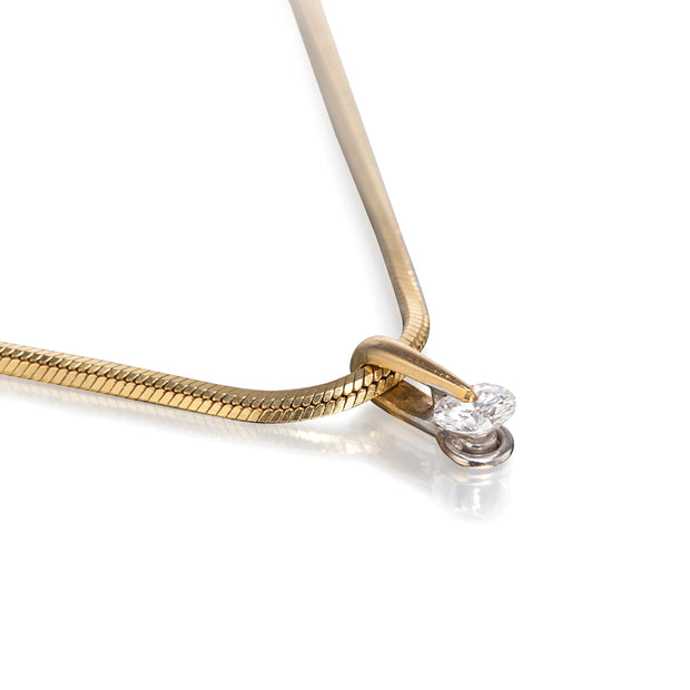 A close-up studio shot of a gold snake chain necklace with a small silver-toned setting holding two clear gemstones, photographed on a white background with a soft reflection.