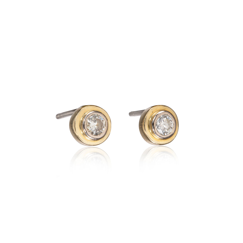 A pair of gold stud earrings with a clear round-cut gemstone in a bezel setting, shown on a white background with a soft reflection below.