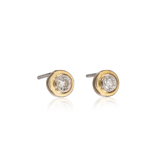 A pair of gold stud earrings with a clear round-cut gemstone in a bezel setting, shown on a white background with a soft reflection below.