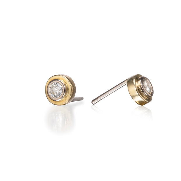 A pair of round gold stud earrings with clear gemstones, shown from two angles on a reflective white background.