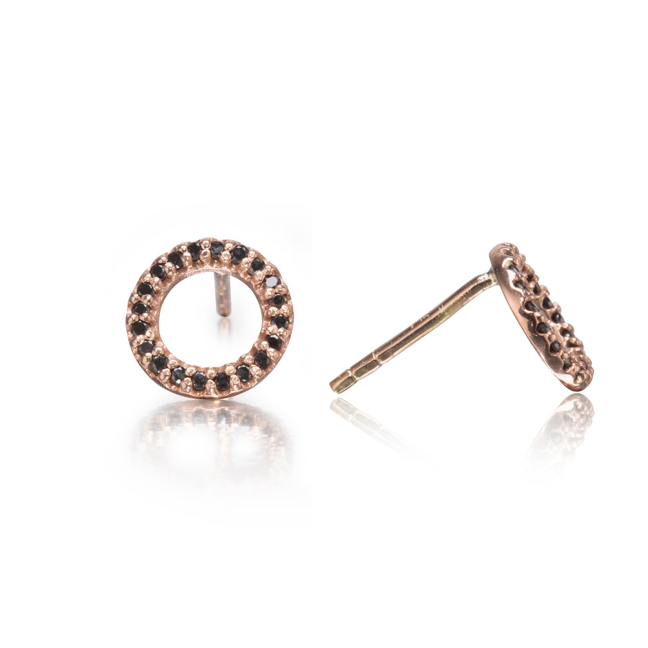 A pair of rose gold stud earrings with a circular halo design set with small black stones, shown on a reflective white background.