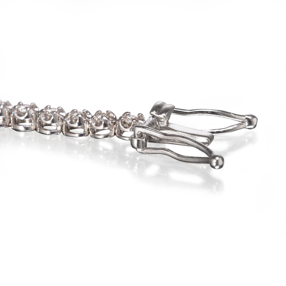 Close-up of a white gold tennis bracelet showing the clasp and part of the chain on a white reflective surface.