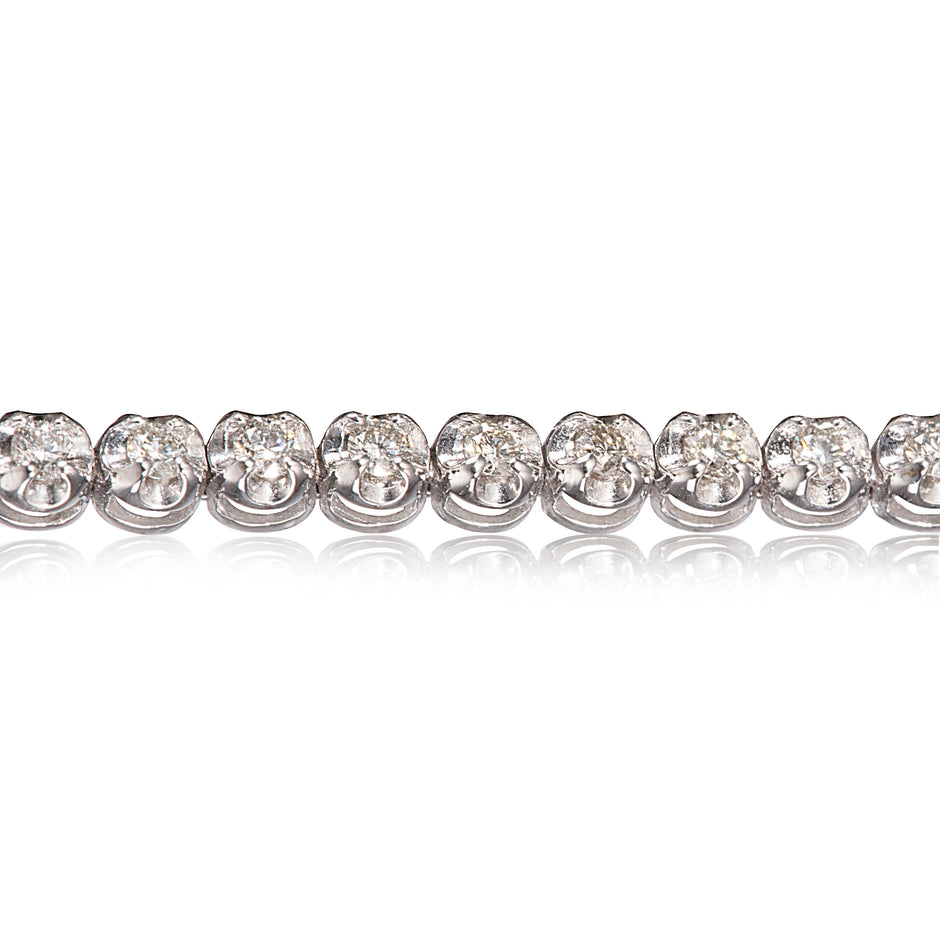 A close-up of a diamond tennis bracelet on a clean white background with a soft reflection below.